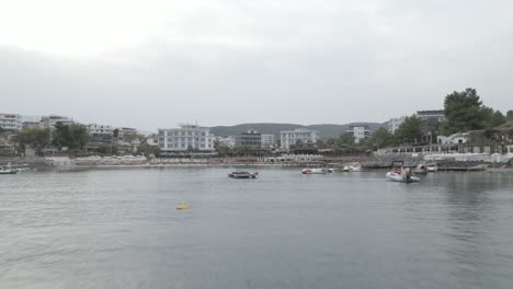 Drone-shot-flying-over-Ksamil-Albania-in-the-morning-on-a-cloudy-grey-day-with-haze-in-the-sky-and-white-hotels-and-houses-in-the-background-with-mountains-and-the-sea-LOG