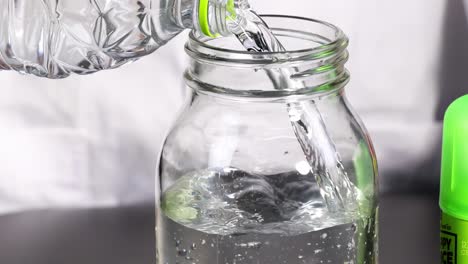 a plastic bottle pours water into a glass jar, with a green cap visible beside it.