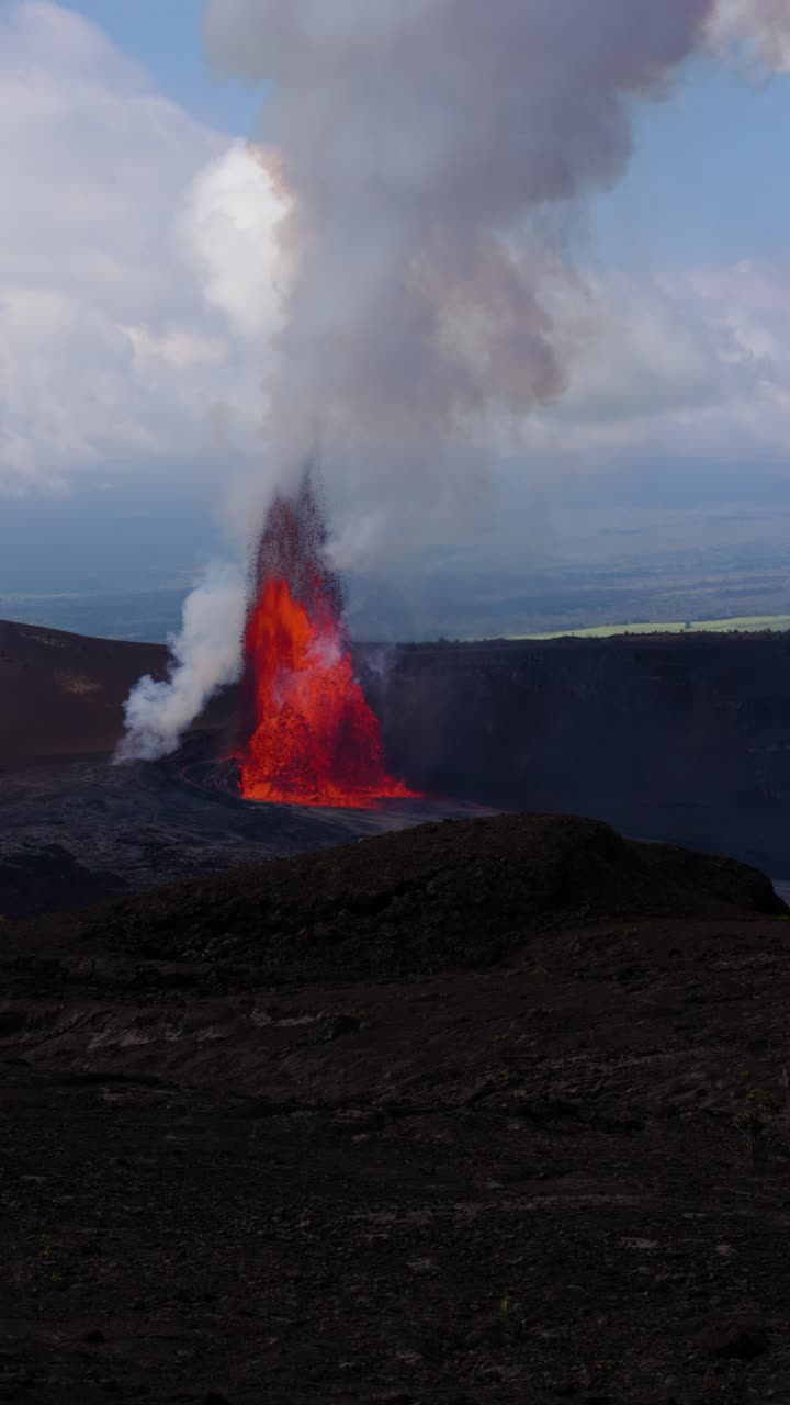 Filmato verticale in slow motion 4K di una colonna di lava isolata che erutta dal cratere Kīlauea, con denso fumo che sale nel cielo coperto sopra un terreno scuro.