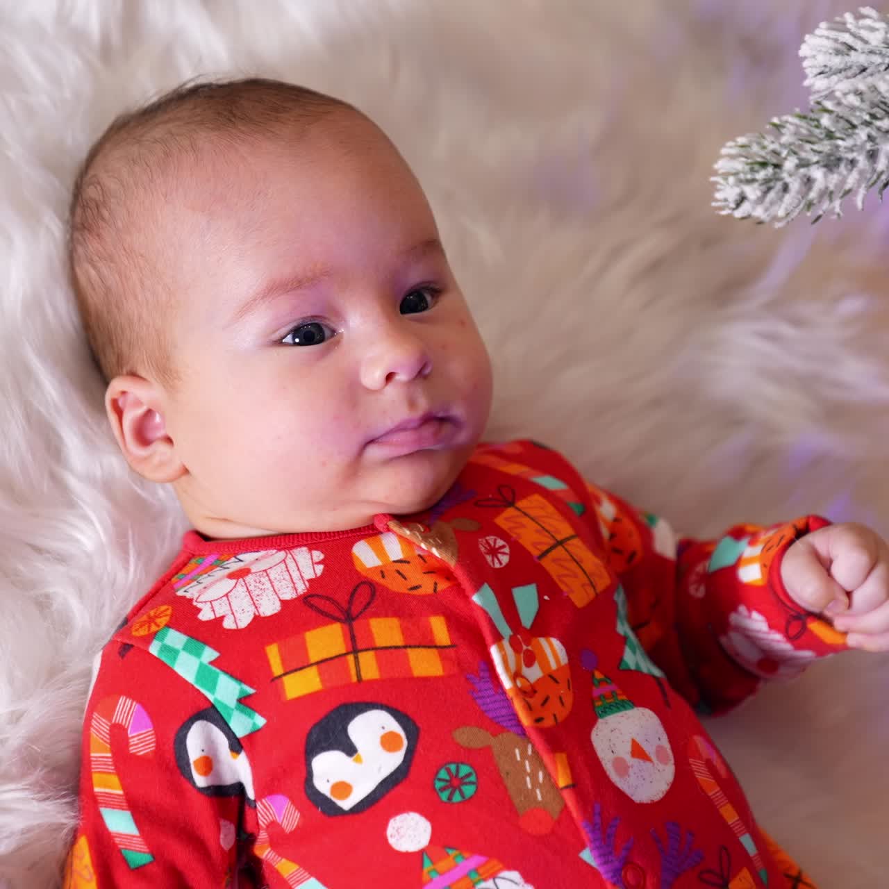 Calm peaceful baby boy in red shirt lies on soft plaid under new year tree. Sweet child looks up at shiny decoration ball hanging over him. View from above