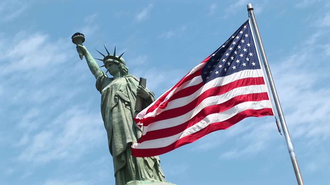 la estatua de la libertad se yergue contra un cielo azul mientras una bandera estadounidense ondea en primer plano