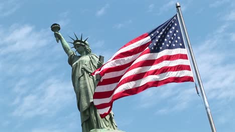 The-Statue-Of-Liberty-Stands-Tall-Against-A-Blue-Sky-As-An-American-Flag-Waves-In-The-Foreground