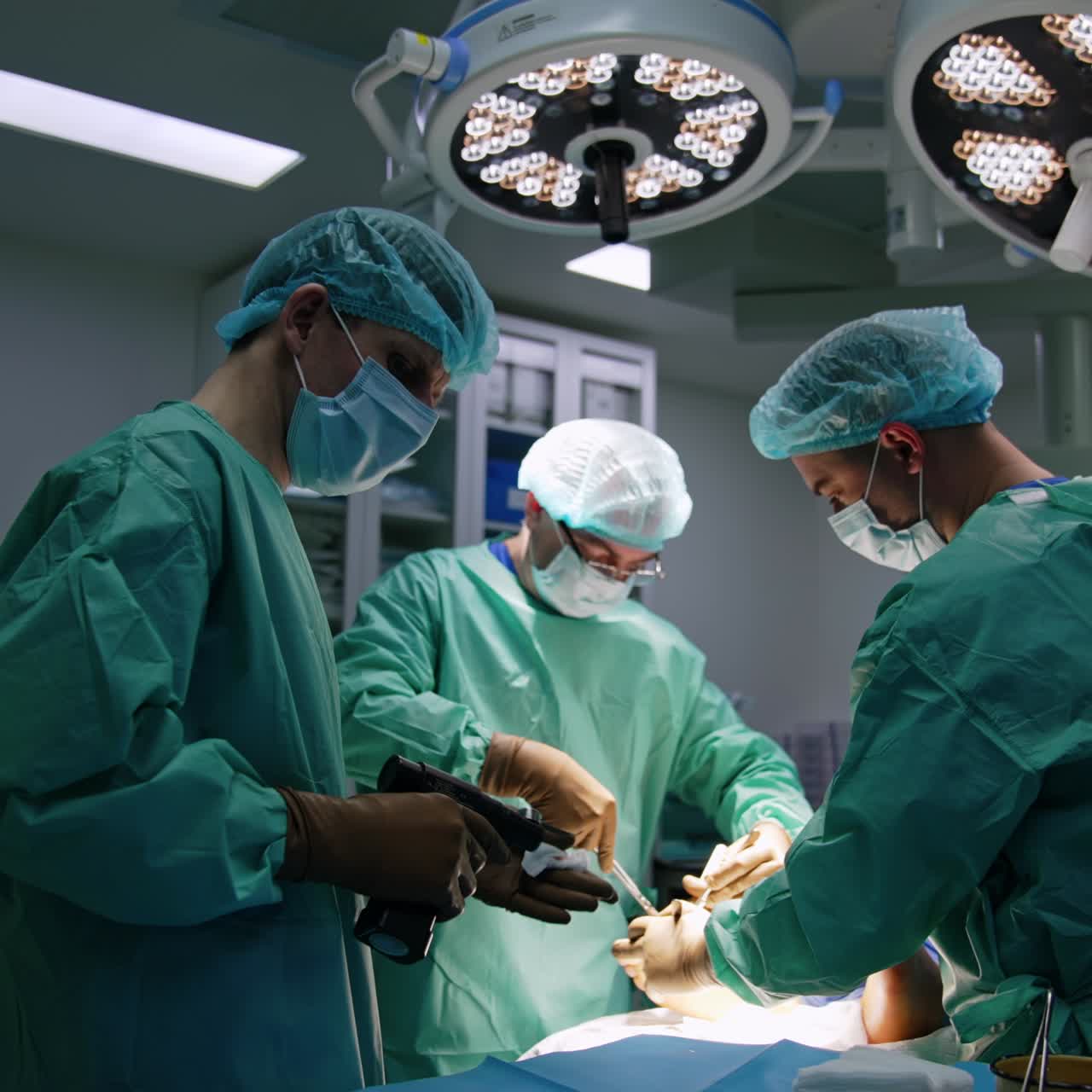 Um auxiliar de médico passa as ferramentas de metal para o cirurgião. Um enfermeiro limpa o dispositivo usado com uma esponja.