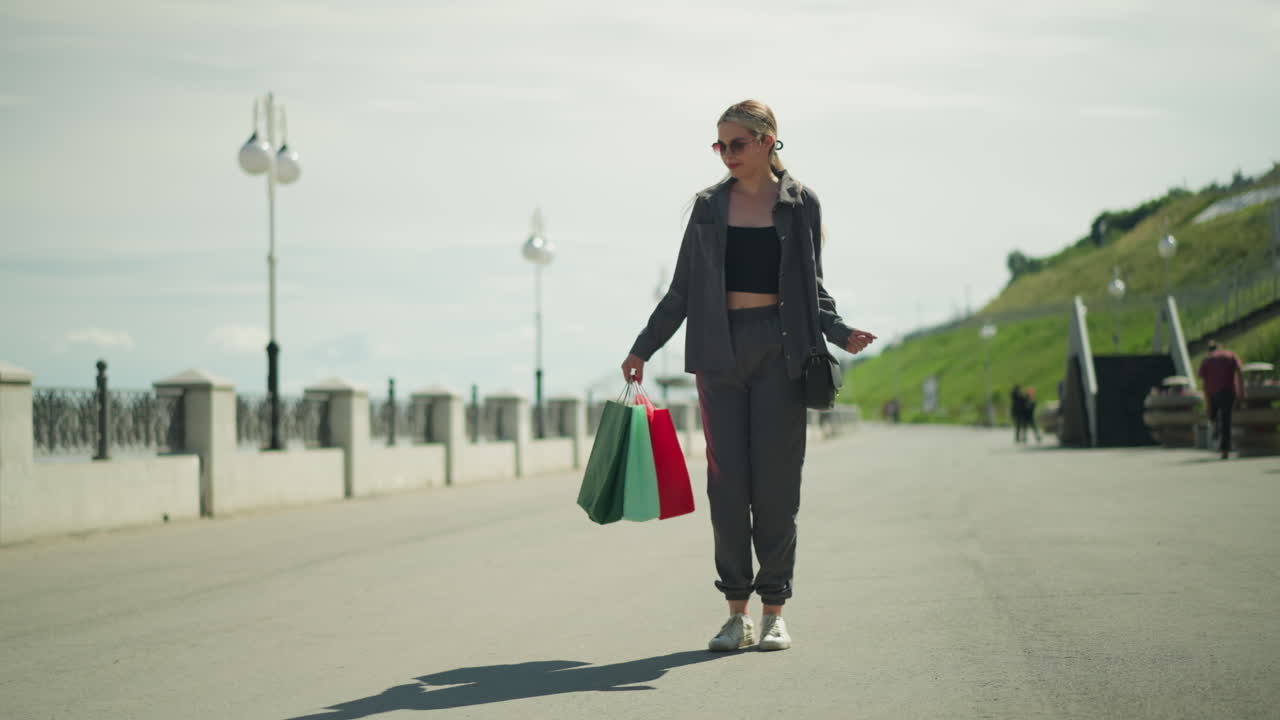 Lady in black crop top under grey jacket dances happily while holding shopping bags in her right hand, her shadow is visible on the ground with a hill on one side and light poles lining the path
