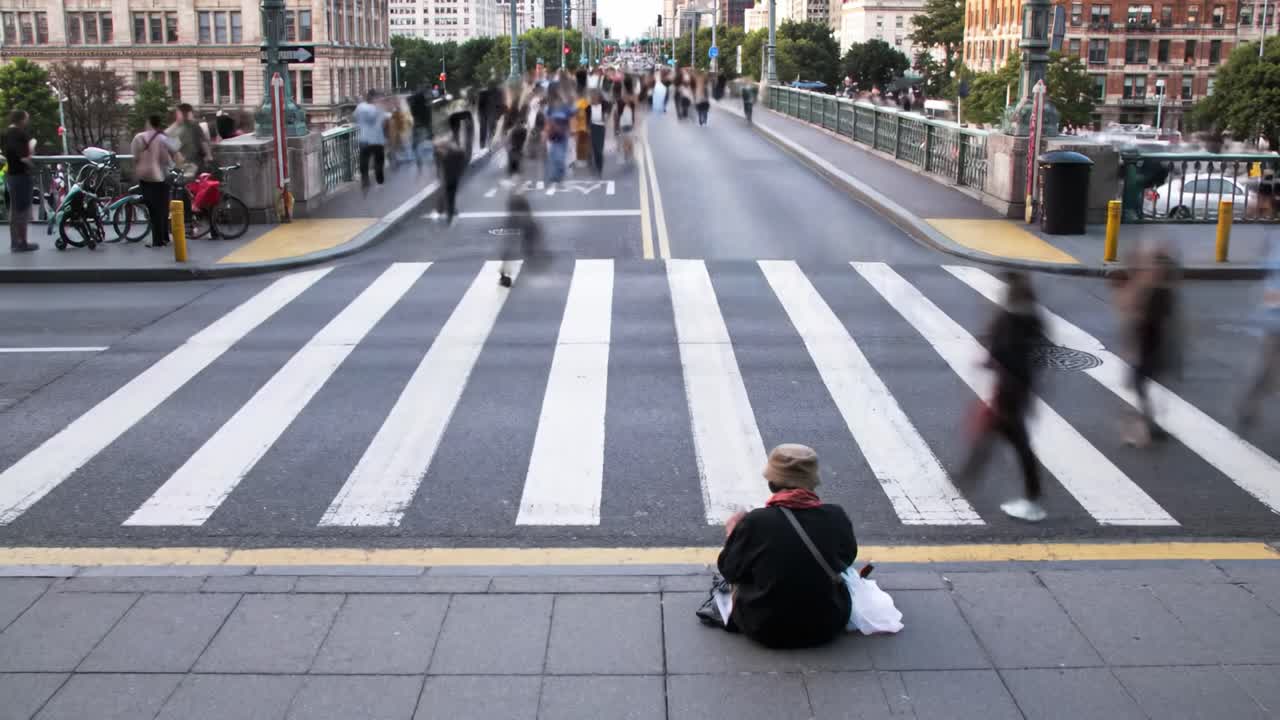 A vibrant urban setting showcases numerous pedestrians crossing a bustling intersection while one individual sits quietly by the curb. The atmosphere reflects the dynamic nature of city life.