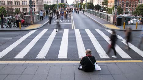 a vibrant urban setting showcases numerous pedestrians crossing a bustling intersection while one individual sits quietly by the curb. the atmosphere reflects the dynamic nature of city life.