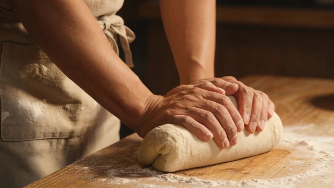 Starting pressing palms, hands rolling dough and forming loaf on floured kitchen counter in apron