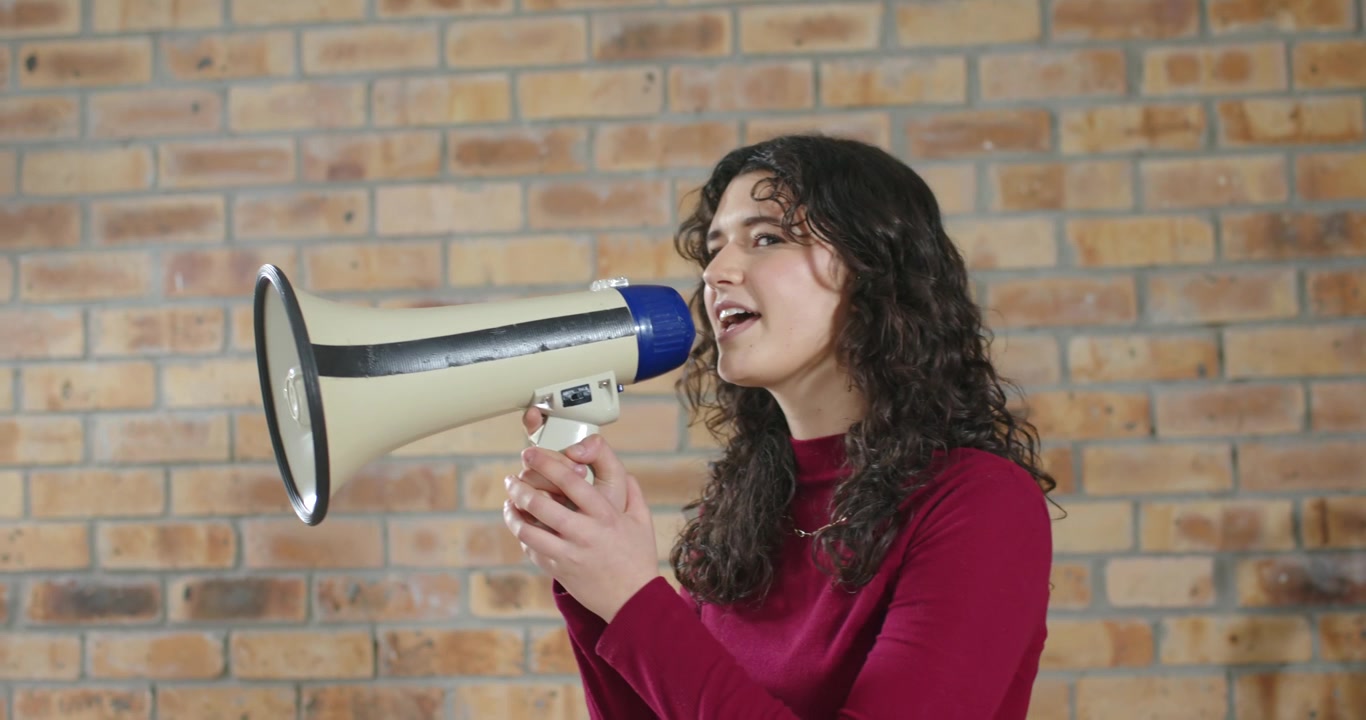 Mujer adulta usando un megáfono para proyectar su voz, ensayando bajarlo y sonriendo ante una pared de ladrillos