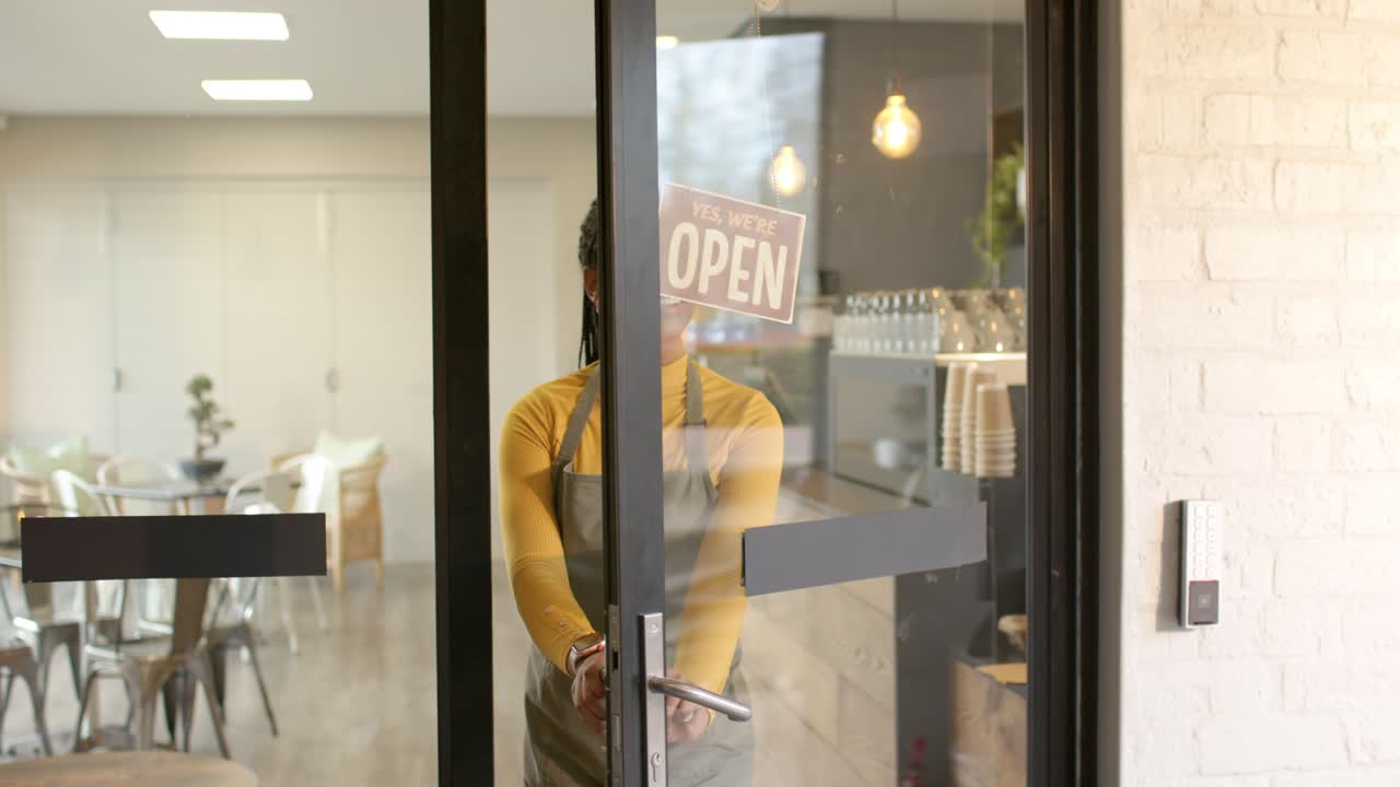 Donna afroamericana che scambia il cartello "chiuso" con "aperto", apre la porta di un caffè e accoglie i clienti.