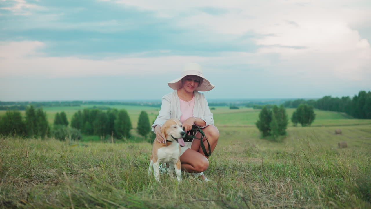 웃는 여성은  모자를 입고 행복한 개 에 앉아서 줄을 잡고 머리를 부드럽게 니다. 배경에는 울창한 초록색 과 나무가 있습니다.