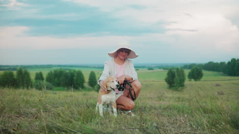 smiling woman in sunhat squatting beside her happy dog, gently tapping its head while holding the leash, background features lush green fields and trees
