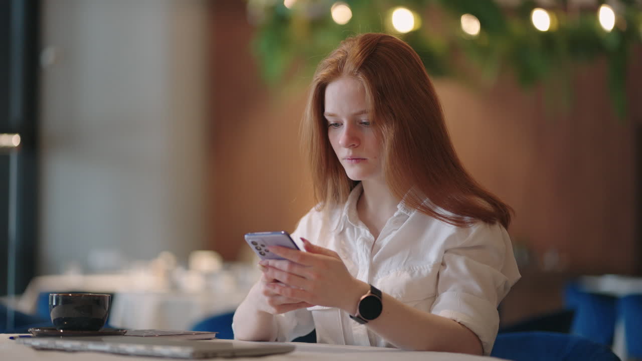 Red-haired Woman working from home using laptop computer while reading text message on mobile phone. Woman using a phone. Serious charming woman using smartphone while working with laptop at home