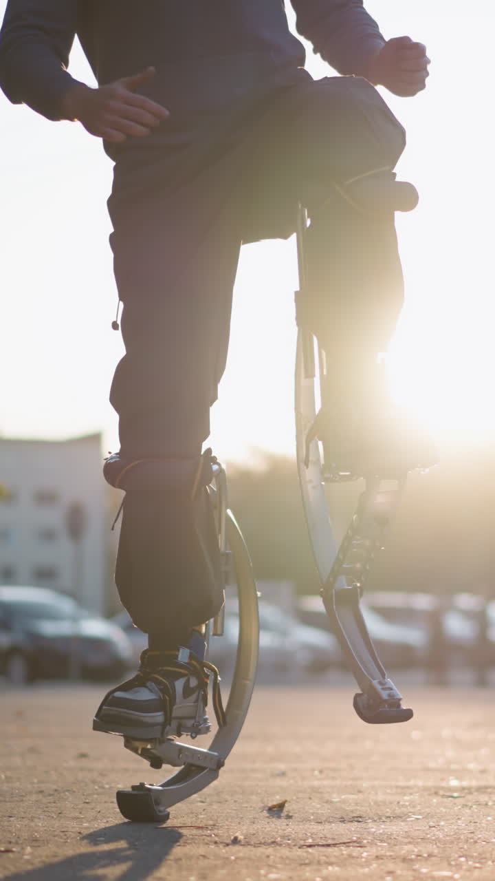 Person with short brown hair wearing gray hoodie and dark gray pants jumping on spring stilts on paved road in residential area surrounded by houses, showcasing unique motion and urban lifestyle