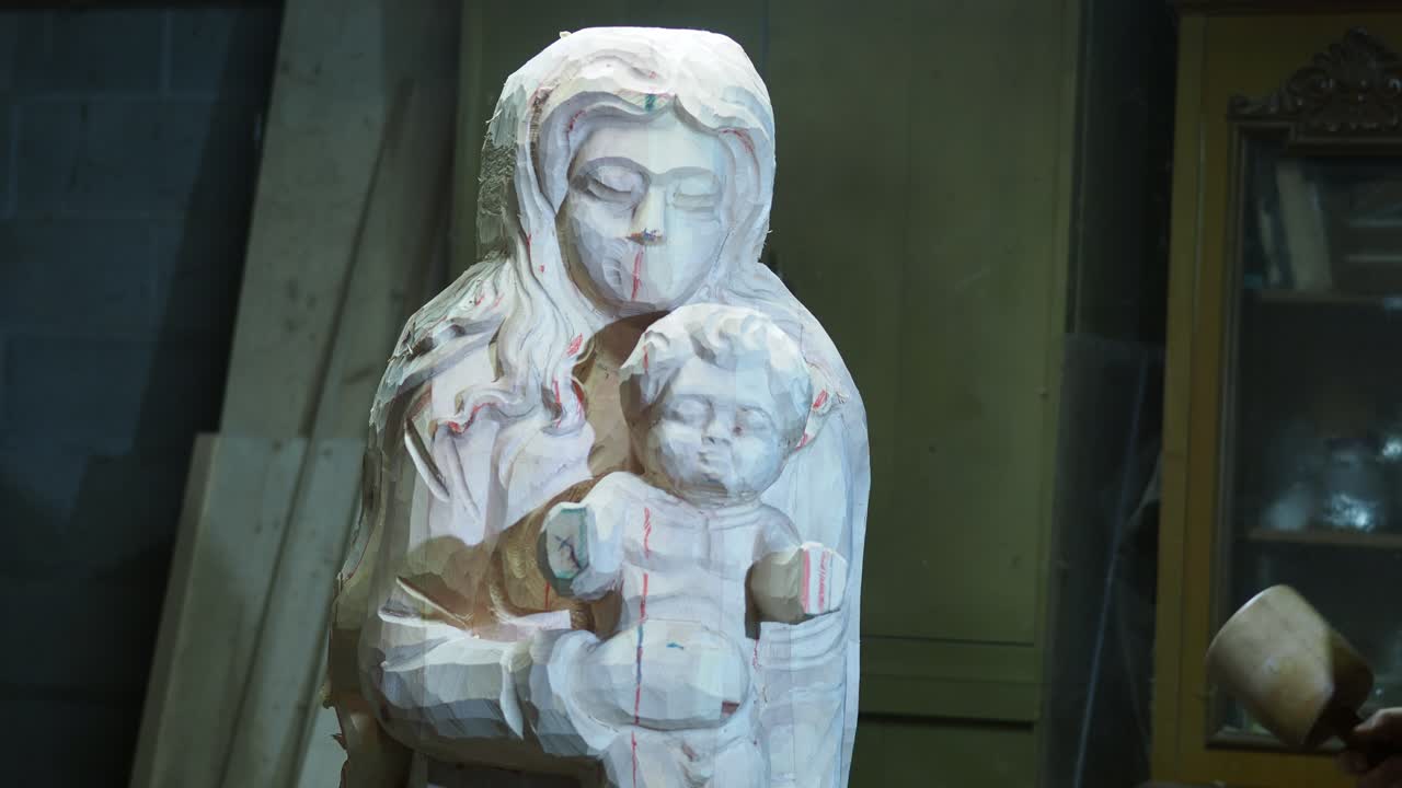 A sculptor stands with a mallet, carefully studying a partially carved wooden Madonna and Child statue, with red guide lines visible, in a traditional workshop setting