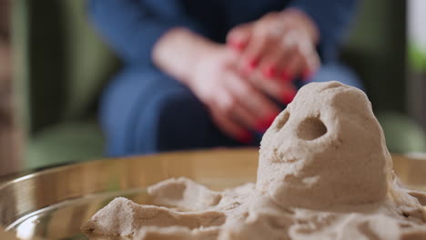 zoomed in view of detailed octopus figure sculpted from sand in metal tray, with blurred figure in blue gown seated in background, showcasing tactile therapy and mindful sculpting activity