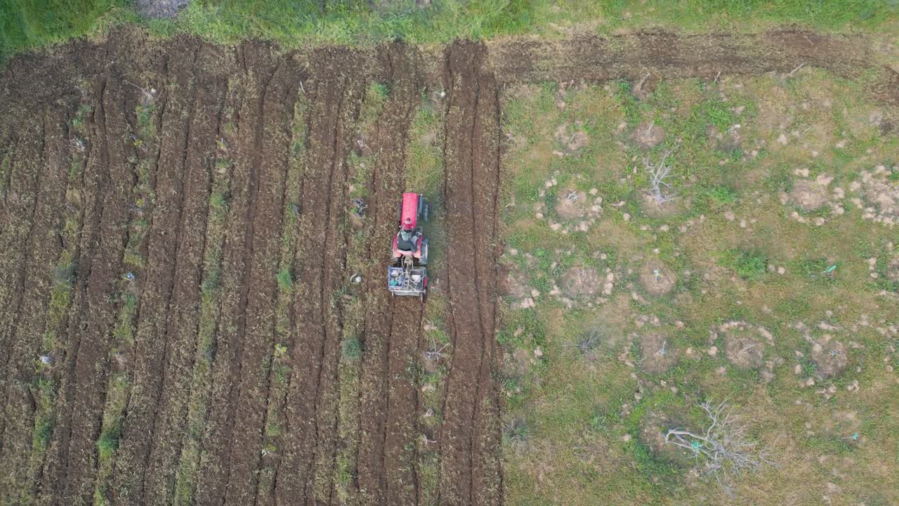 Cinematic top-down drone shot of a small tractor plowing a tree-lined field. One section is fully tilled, and the tractor moves into position to begin a new row.