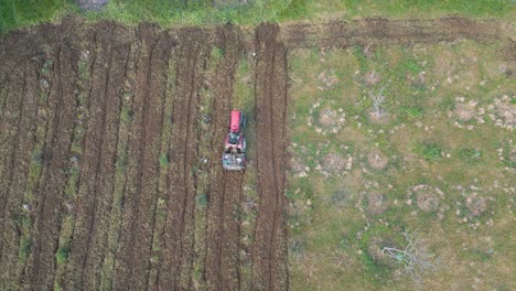 cinematic top-down drone shot of a small tractor plowing a tree-lined field. one section is fully tilled, and the tractor moves into position to begin a new row.