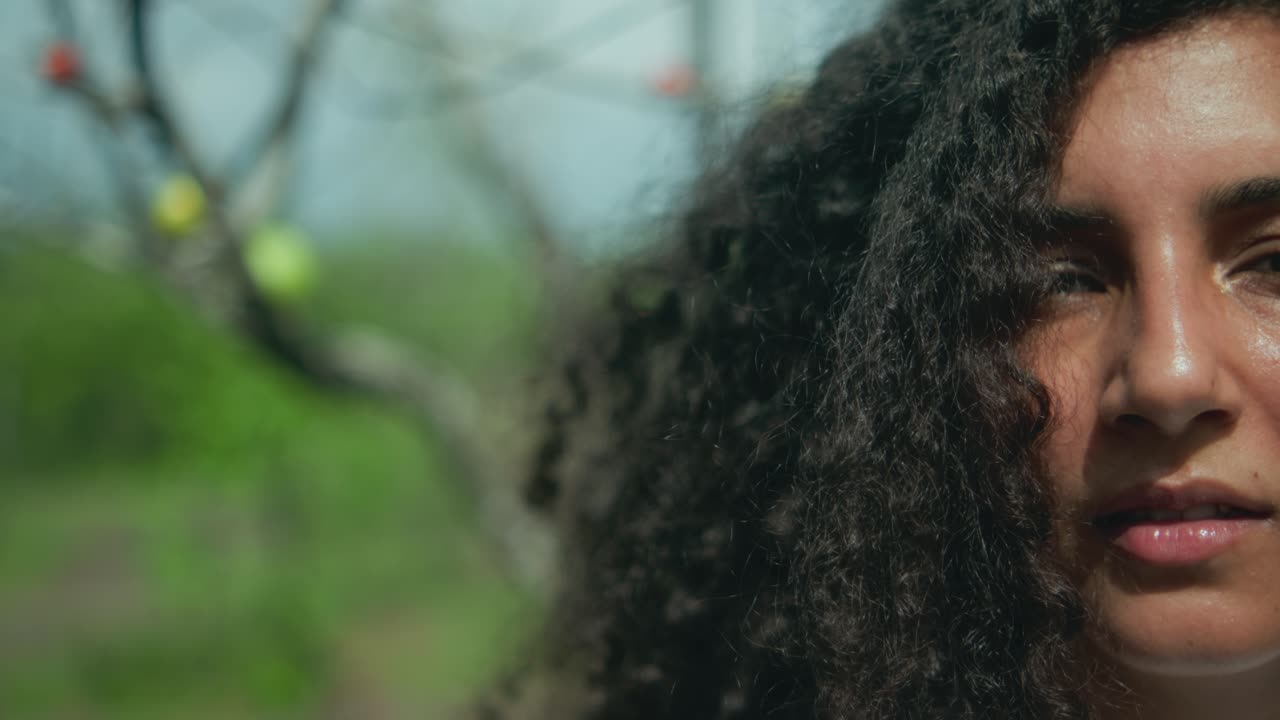 Tight close-up of a Middle Eastern woman’s face and voluminous curly hair in sunlight, with a defocused tree bearing colorful fruit in the background, capturing texture, mystery, and natural presence