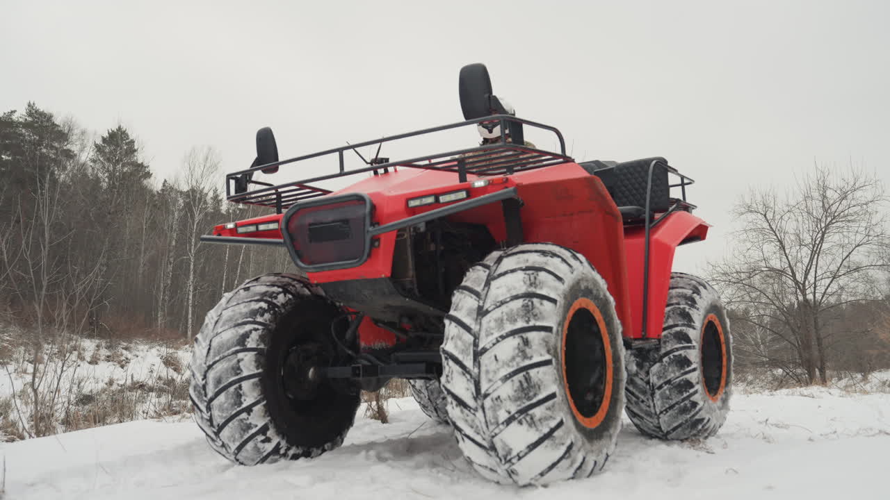 Rød ATV parkeret på snedækket mark, overdimensionerede flotationdæk med snedækket slidbane, frontvinkel viser bagagebærer og forlygte, fører i hjelm og camouflagejakke siddende bag styret, motor i tomgang.