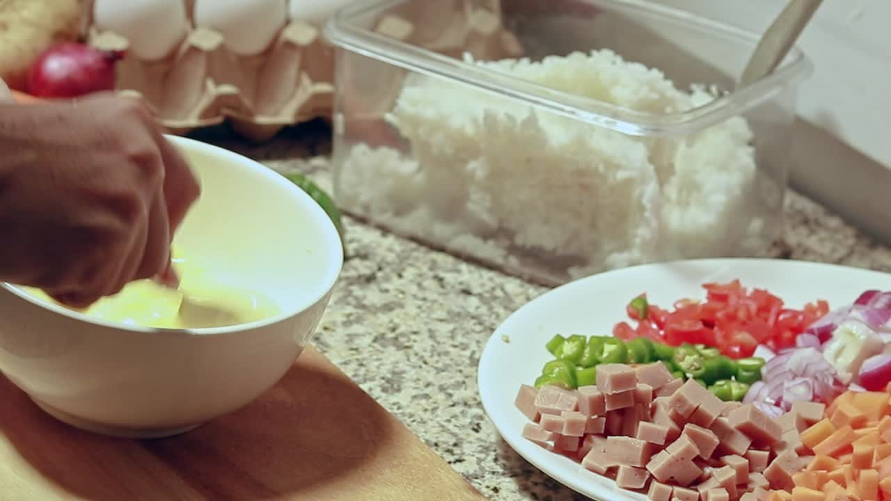 A kitchen scene with eggs being beaten, rice in a container, and diced vegetables and ham on a plate, ready for cooking fried rice