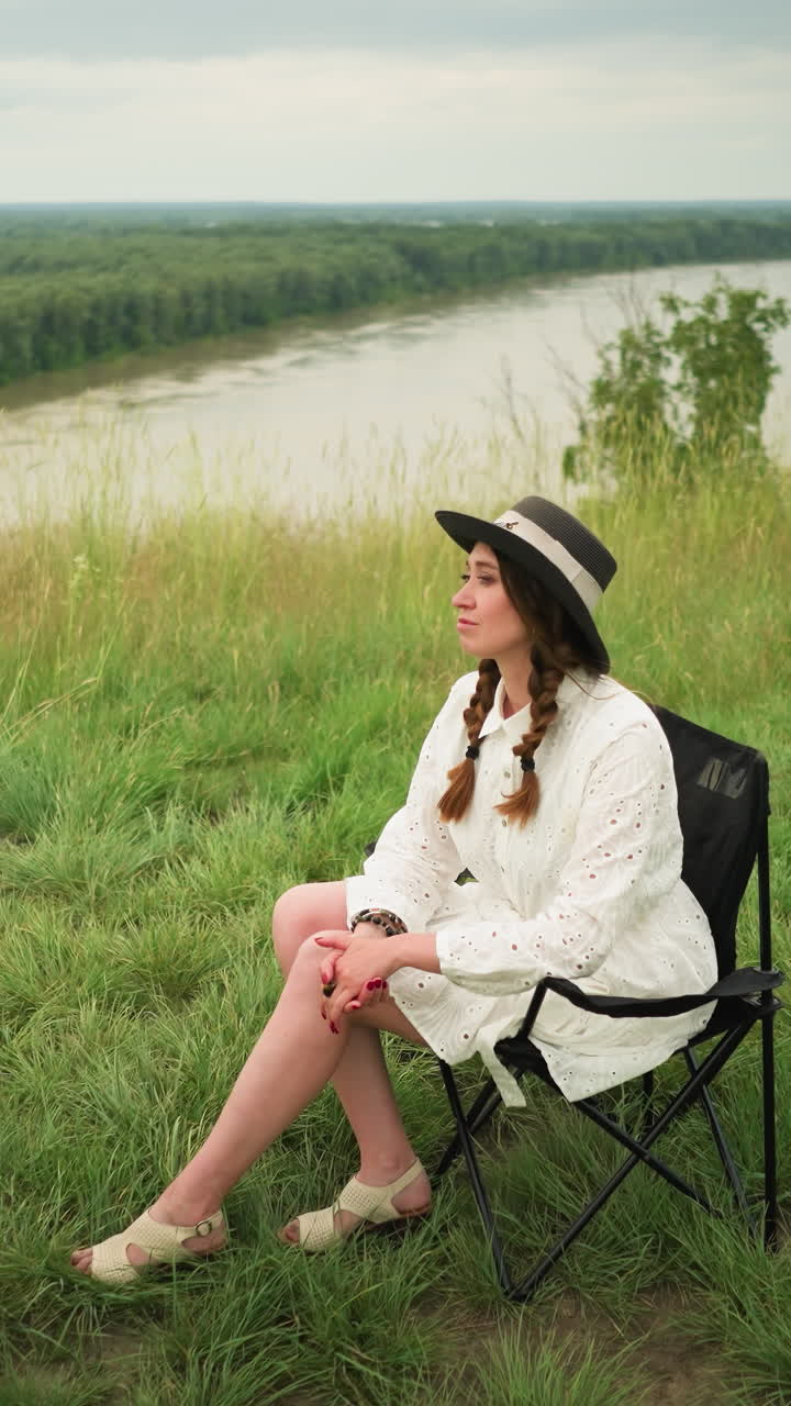 A cute woman wearing a stylish hat and a white dress is seated on a chair in a lush grass field beside a tranquil lake. She poses elegantly with a serene expression under a cloudy sky