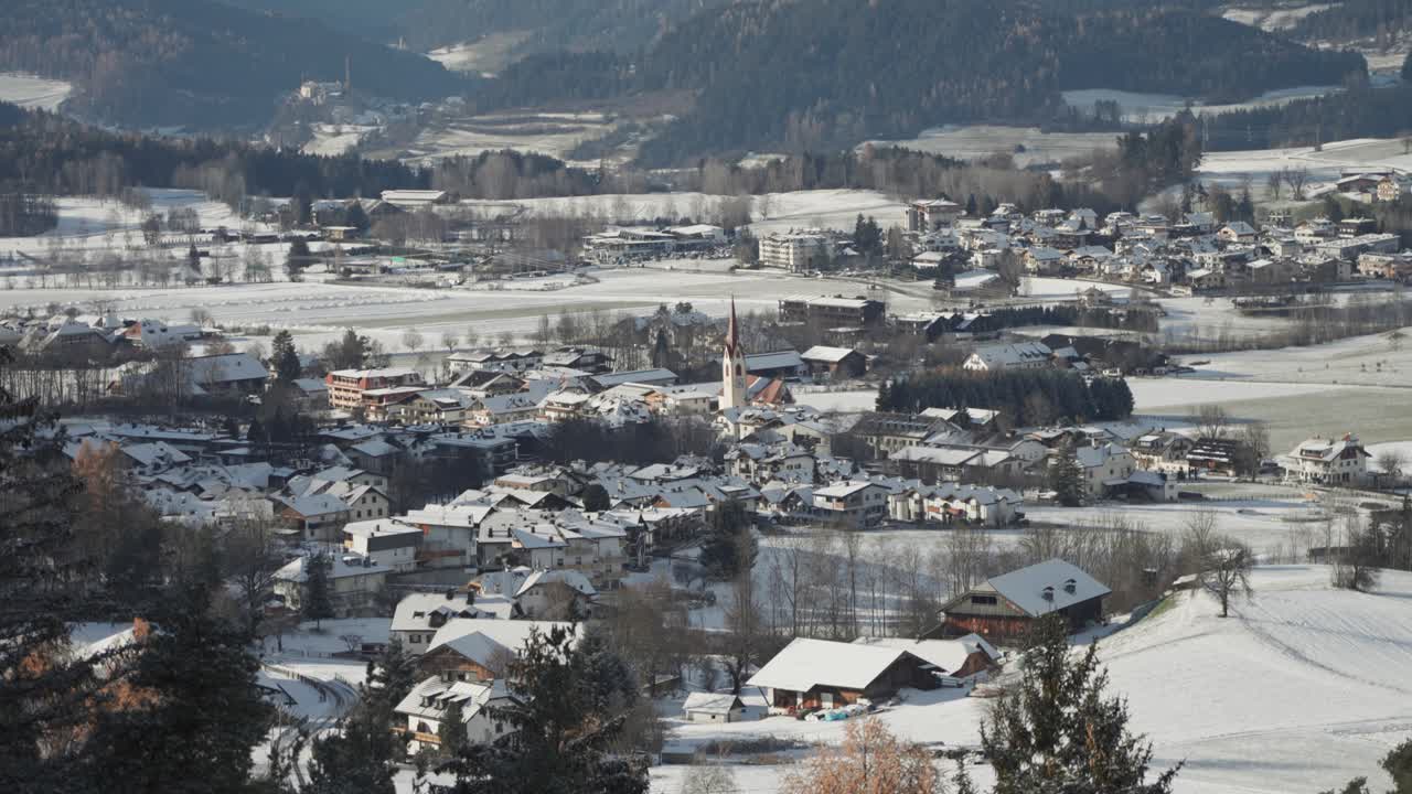 A snow-covered village with the red-steepled church in the center at the foot of forested hills in South Tirol, Italy.