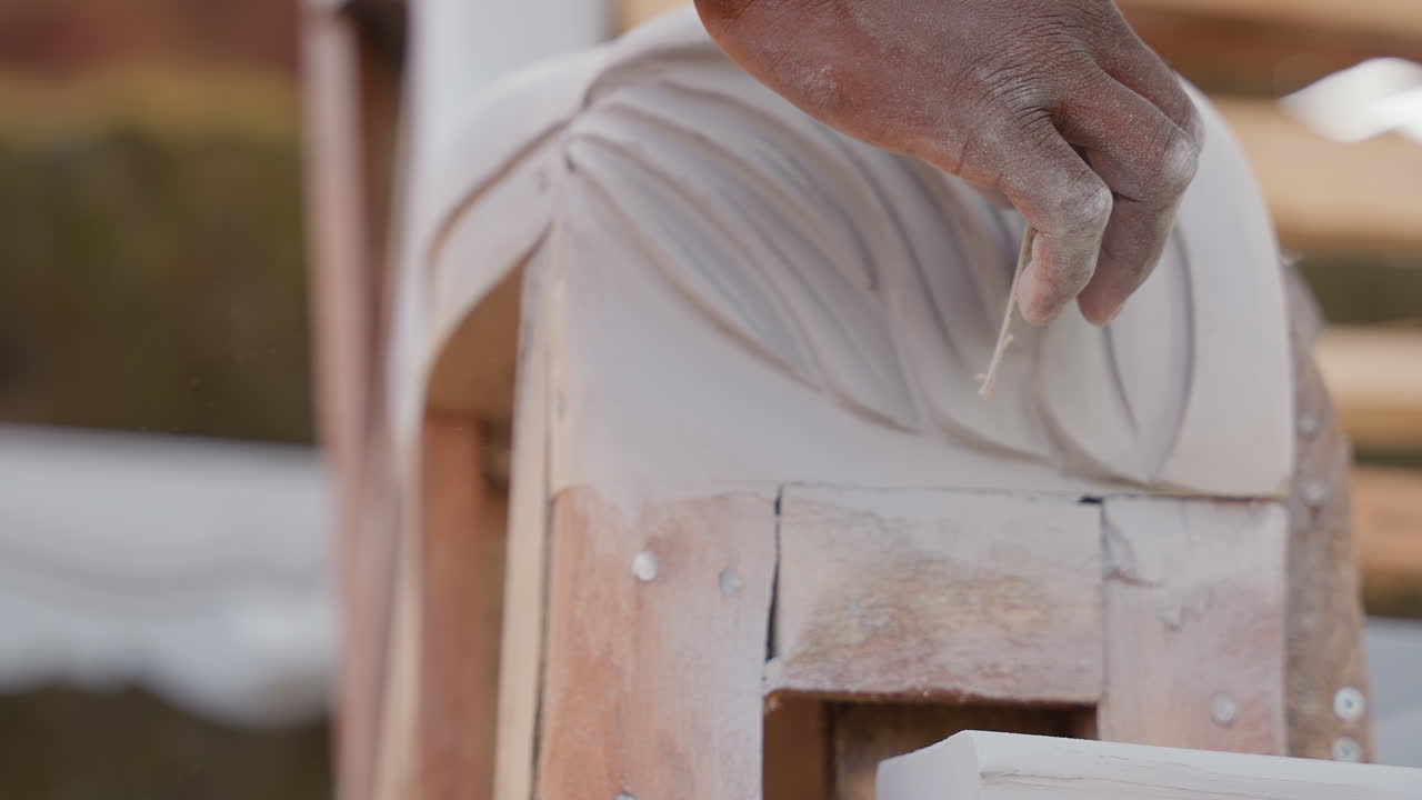 Primo piano di mani che intagliano modanature in legno con scalpello e polvere di levigatura, atmosfera di laboratorio di restauro, artigiano che modella un rilievo decorativo a foglia, precisione attenta e tecnica vintage, luce naturale calda.
