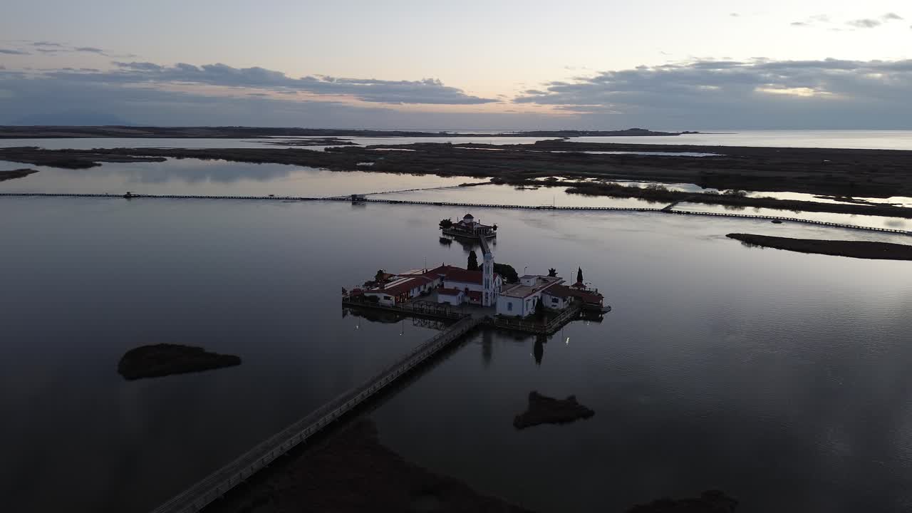 Filmisk luftfoto af Sankt Nikolaj Kloster, bygget på en lagune i Porto Lagos, Grækenland, med rolige vandrefleksioner og en træbro under en fredfyldt blå time ved solnedgang.