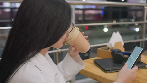 close up side view of woman in glasses sipping coffee while texting on phone, seated by wooden table with napkin holder and headphones, night traffic bokeh seen through metal railing and glass panel