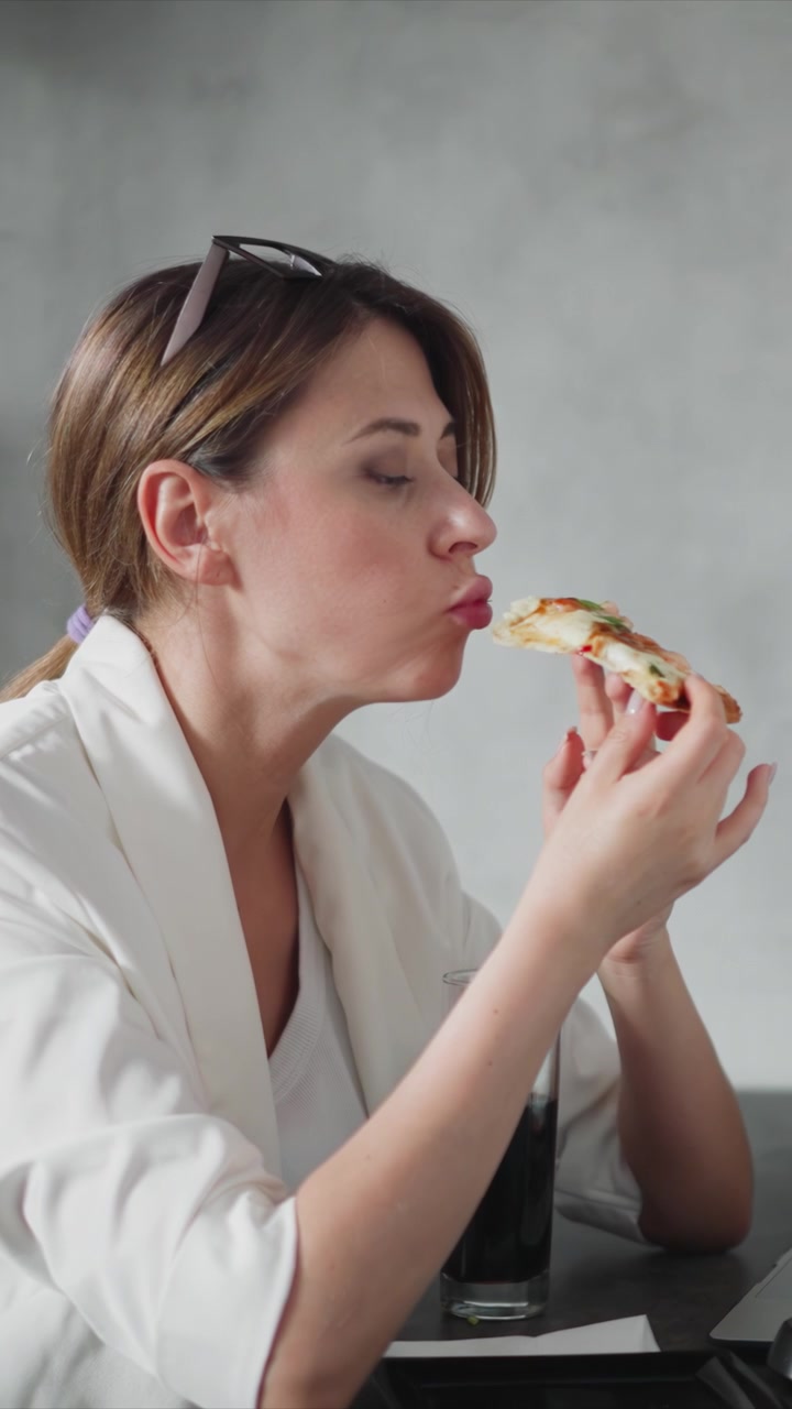 Close up of woman eating pizza while seated at table with glass and laptop. Woman holds slice takes bites against plain gray background. Soft lighting highlights her actions and surroundings