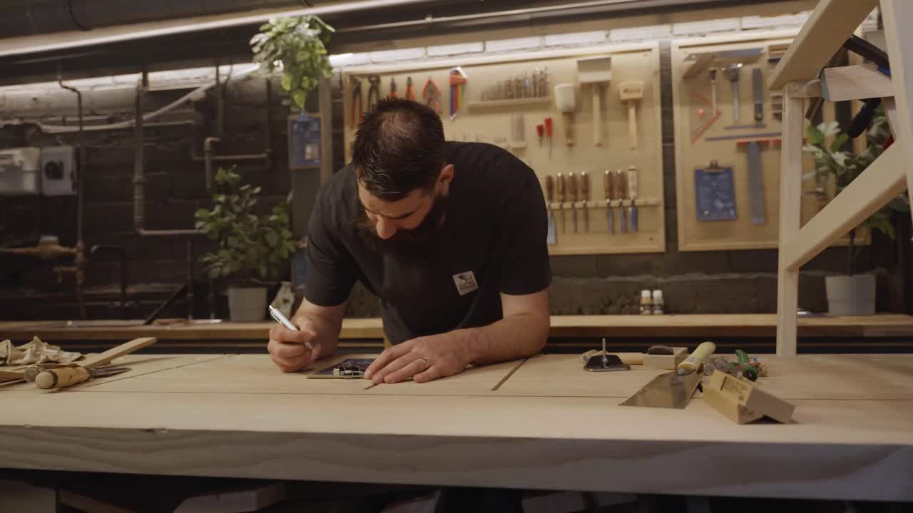 A craftsman leans over a wooden workbench in a modern workshop, surrounded by tools and materials, concentrating on his woodworking project and design details