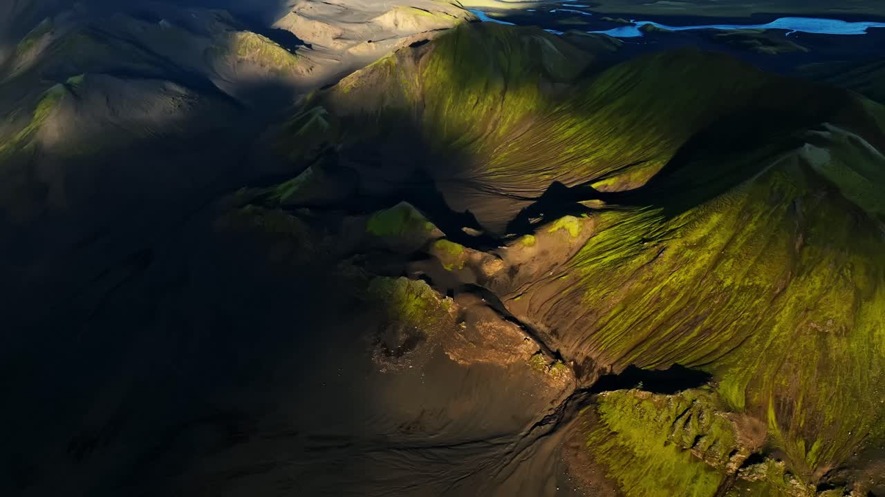 Aerial pan-up over Iceland’s volcanic highlands, lifting from shadowed craters to reveal bright moss ridges, winding lakes and vast rugged terrain under crisp warm sun light