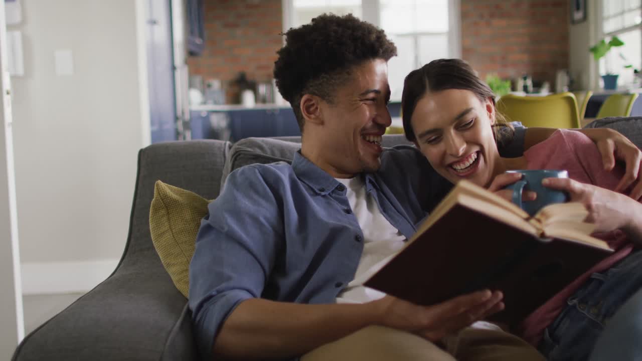 Happy biracial couple sitting on sofa in living room, reading book and drinking coffee