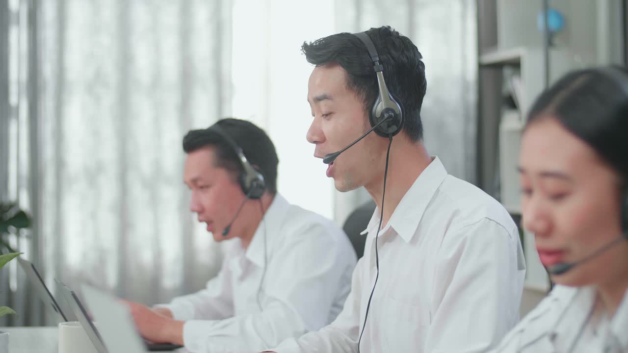 Three Asian Call Center Agents Wearing Headsets Sitting On The Chairs And Typing On Computers While Speaking To Customers On The Call At The Office