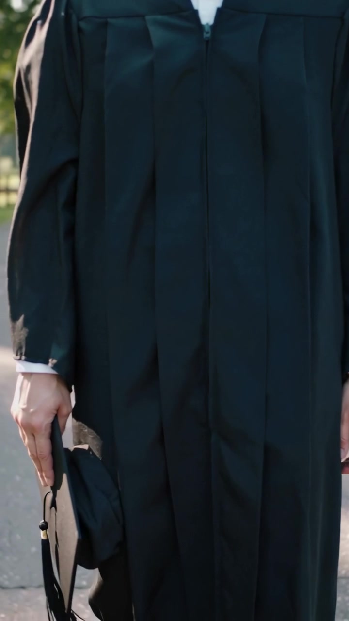 Video verticale: Laureato in toga in piedi che avverte un leggero ondeggiamento e si sistema il tocco su un sentiero del campus, in posa.