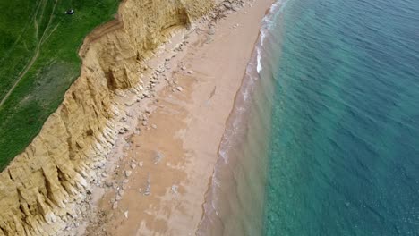 drone video aimed down over a jurassic coast beach in dorset. the grass top cliffs on the left of the shot, beach with large rocks on the middle and the clear blue ocean on the right.