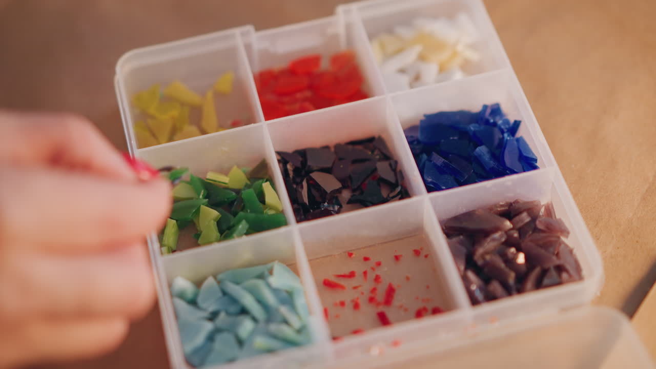 Primer plano de bandeja con coloridas teselas de mosaico mostrando manos clasificando piezas para decoración de regalos, luz ambiental cálida, superficie de mesa de madera, compartimentos organizados con fragmentos azules, rojos, verdes y amarillos, cuidadoso