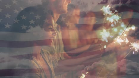 Animation-of-american-flag-waving-over-group-of-friends-holding-sparklers-on-beach