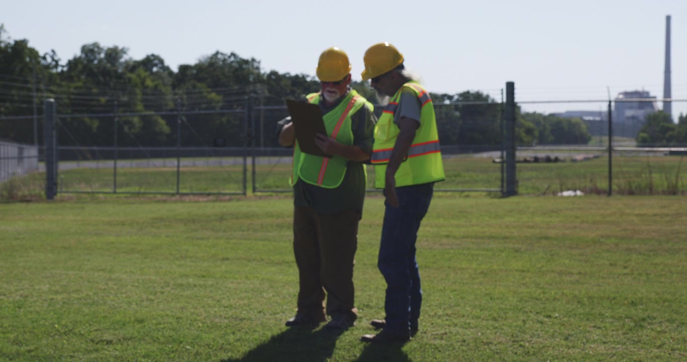 Two industrial workers discuss site conditions on grass, one holding clipboard near facility