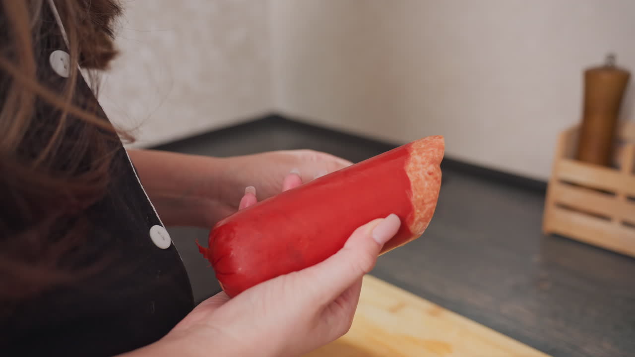 close up of person peeling plastic cover from hotdog with fingers during food preparation process in kitchen standing beside dark wooden countertop with cutting board and stove visible in background