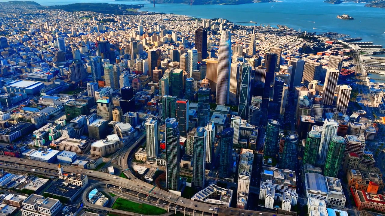 Panorama do horizonte de São Francisco com autoestrada e baía ao fundo. Vista aérea de malha urbana densa, aglomerado de torres, rampa de autoestrada curva e estreito de Golden Gate distante sob céu azul.
