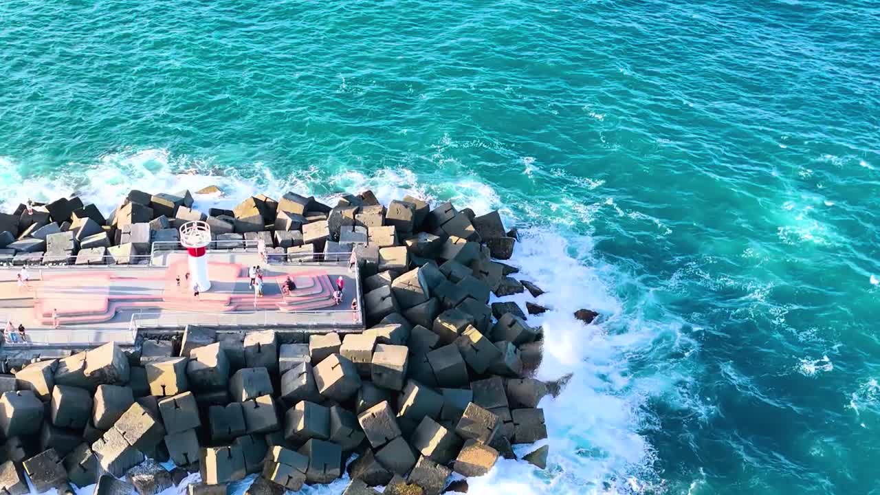 Drone footage of waves crashing against a rock jetty in clear blue waters.