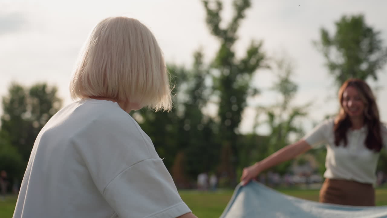 two girlfriends spreading picnic blanket on lush green grass in sunny park with blurred trees in background, joyful outdoor gathering scene showing cooperative gesture and warm summer friendship mood