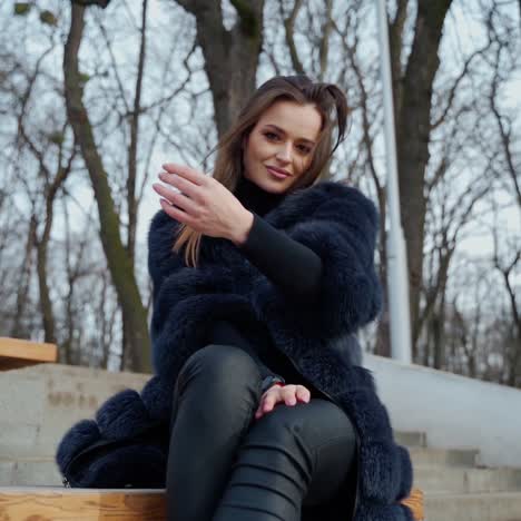 beautiful girl in a coat sitting on a bench in a city park. portrait of a luxury young woman in fur coat looking at camera and beckon with hands to herself. slow motion.