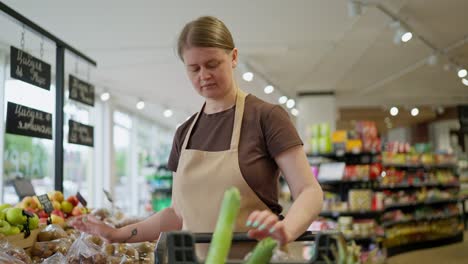 A-confident-brunette-girl-with-blue-eyes-in-a-brown-T-shirt-and-a-light-brown-apron-puts-goods-on-the-counter-from-her-wheelbarrow-during-a-work-shift-in-a-supermarket