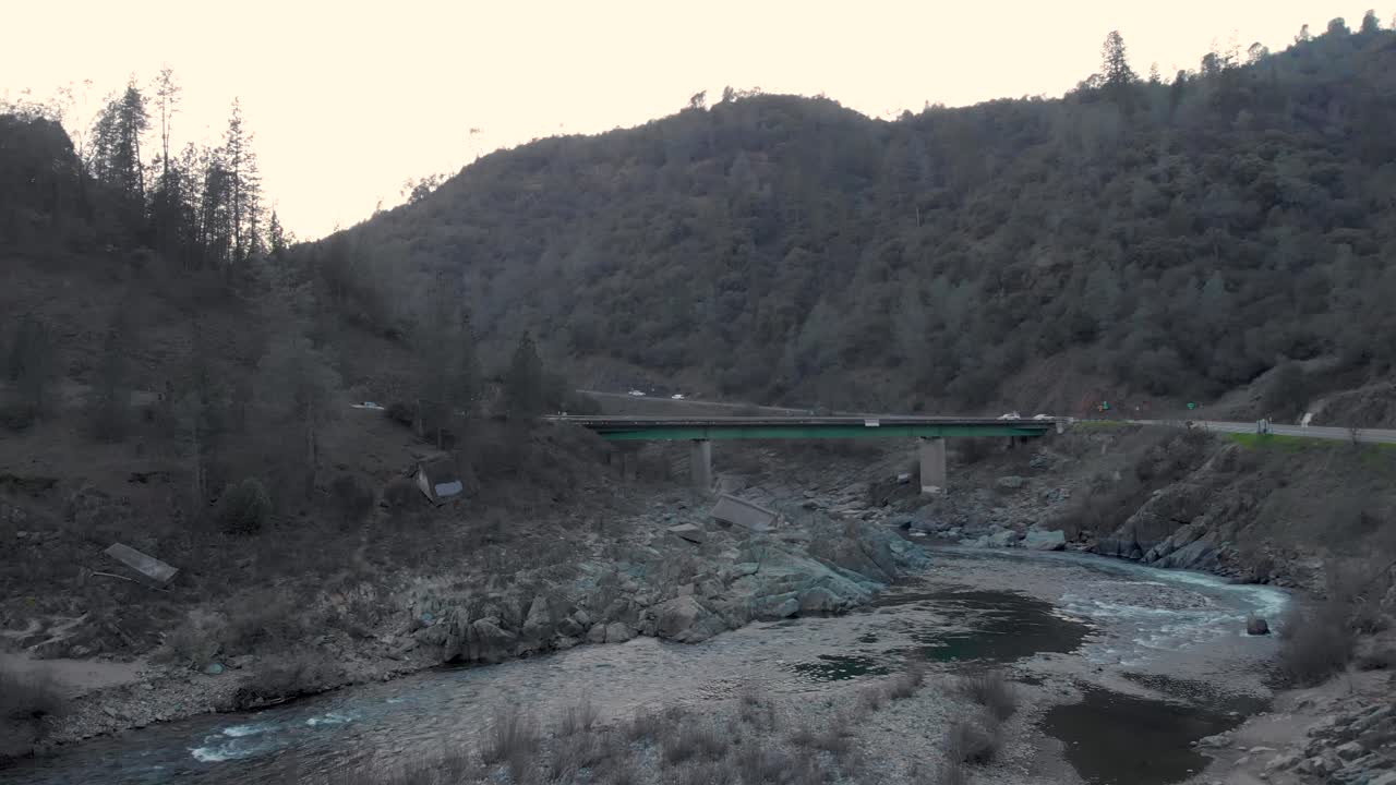 캘리포니아 주 오번에 있는 아메리칸 리버의 녹색 다리를 향해 물과 바위 위를 낮게 날아다니는 드론 - 녹색 나무와 산으로 둘러싸여