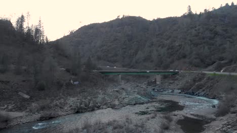 Drone-flying-low-over-water-and-rocks-towards-a-green-bridge-in-the-American-River-in-Auburn,-California---surrounded-by-green-trees-and-mountains