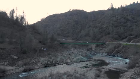drone volando bajo sobre el agua y las rocas hacia un puente verde en el río americano en auburn, california - rodeado de árboles verdes y montañas