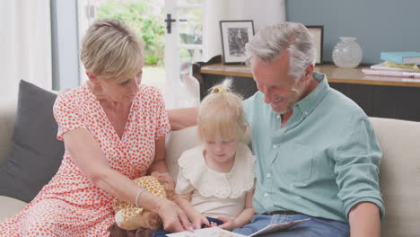 grandparents sitting on sofa with granddaughter at home reading book together