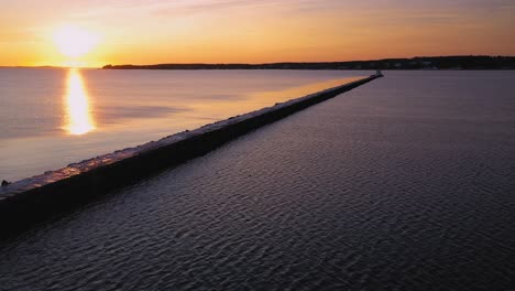 Aerial-footage-flying-backwards-at-sunrise-on-the-Maine-coast-away-from-a-long-pier-with-a-lighthouse-at-the-end-of-it